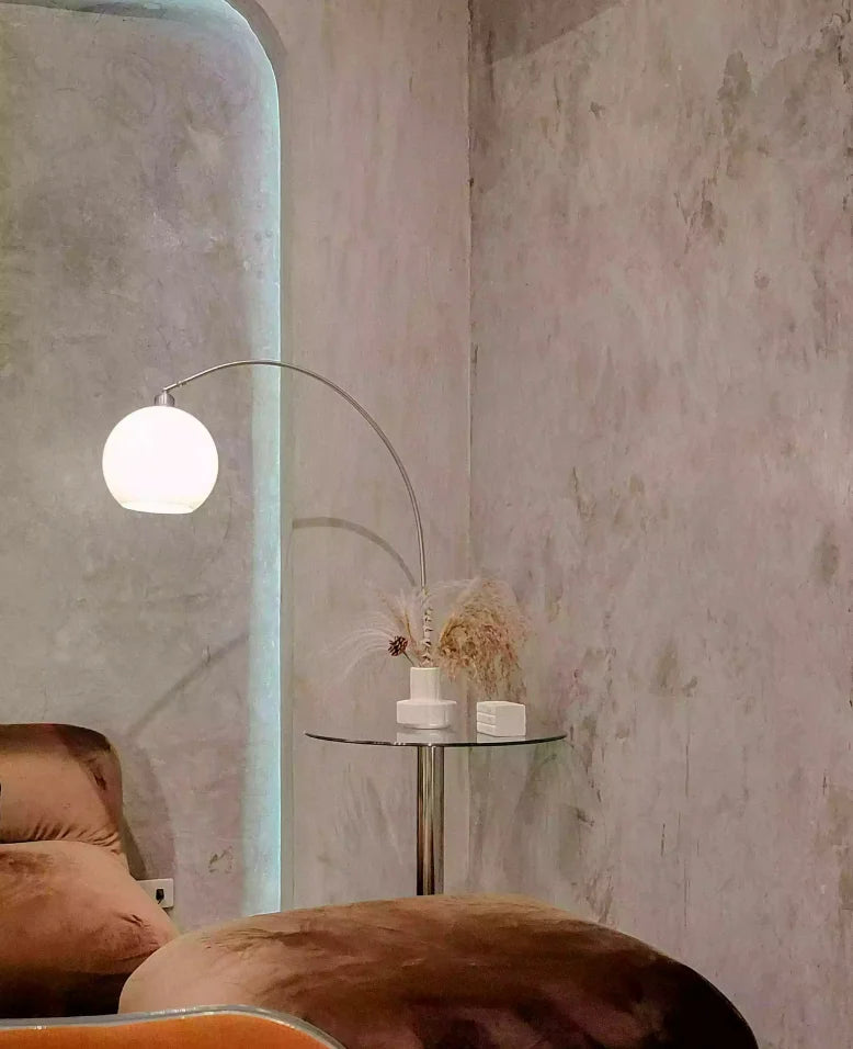 Intérieur design moderne avec à gauche une lampe sur pied à globe lumineux près d’un mur en béton brut et à droite un salon au style minimaliste avec un canapé arrondi crème, une étagère murale organique rétro-éclairée et des objets décoratifs en céramique.