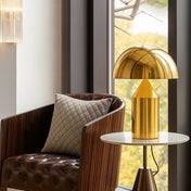 Lampe champignon dorée MORELLO posée sur une table d’appoint en marbre, à côté d’un fauteuil en cuir capitonné et bois foncé avec coussin matelassé, dans un intérieur lumineux avec vue sur un paysage verdoyant.